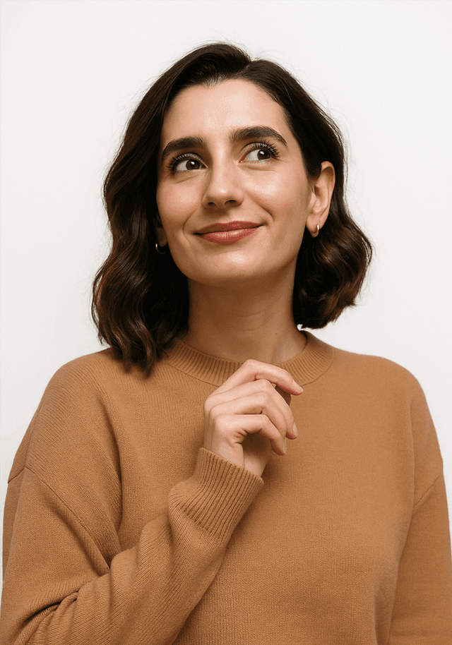 A woman with shoulder-length, wavy dark hair is smiling slightly while looking upwards. She is wearing a cozy, light brown sweater and has a hand raised near her chin, suggesting a thoughtful pose. The background is plain white, which keeps the focus on her face. Her makeup is natural, enhancing her features without being overpowering. She has small hoop earrings that add a touch of elegance to her look.