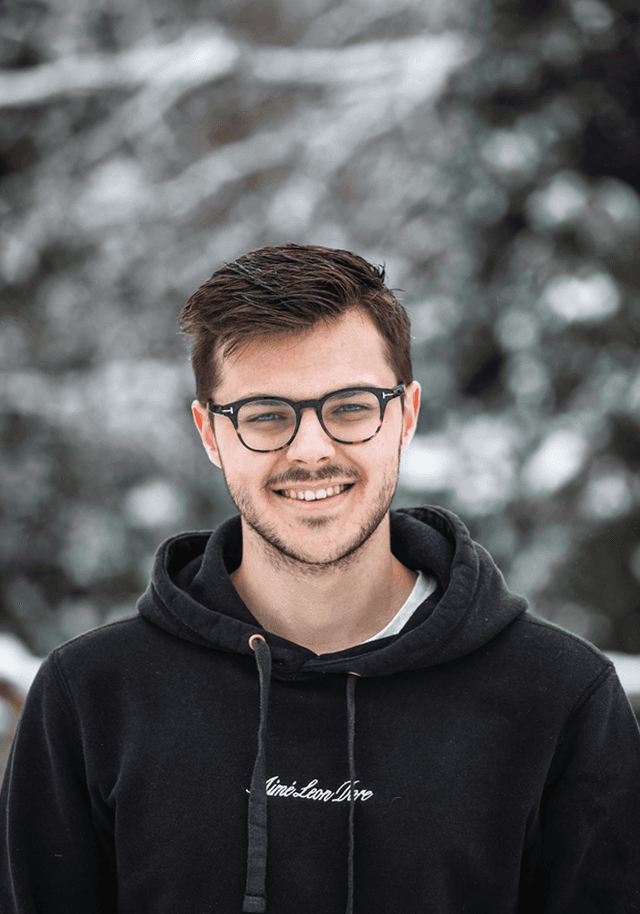 A young man with short, styled hair and glasses is smiling at the camera. He is wearing a black hoodie with a white logo. The background is snowy and blurred, suggesting a winter setting.