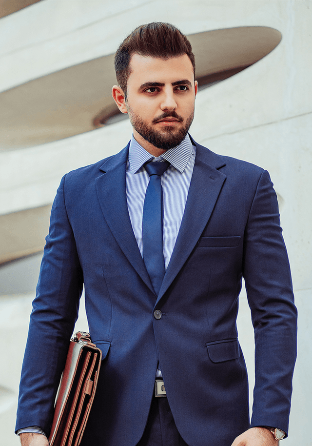 A well-dressed man in a navy suit and a striped shirt, holding a brown leather briefcase. He has a well-groomed beard and styled hair, standing confidently against a modern architectural background.