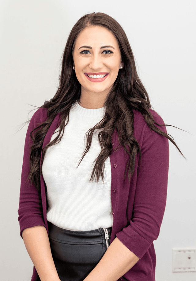 A woman with long, dark hair styled in loose waves, smiling brightly. She is wearing a white sweater and a maroon cardigan, paired with a black skirt. The background is plain white, giving a clean and professional look. Her makeup is natural, enhancing her features, and she has a friendly demeanor.